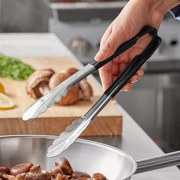 A person using Vollrath Jacob's Pride stainless steel scalloped tongs to pick up mushrooms from a pan.