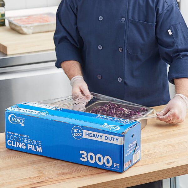 A person in gloves uses Choice Heavy-Duty Foodservice Film to cover a blue container of food.