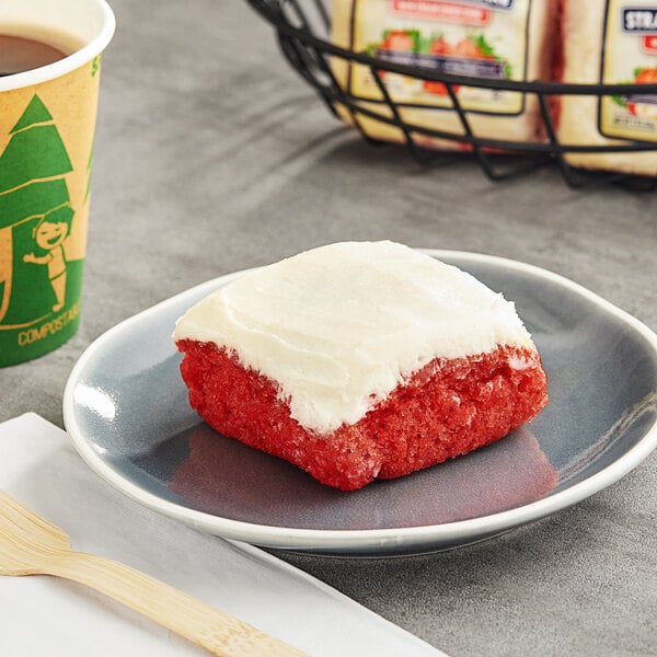 A Ne-Mo's Bakery strawberry cake square with white frosting on a plate next to a cup of coffee.