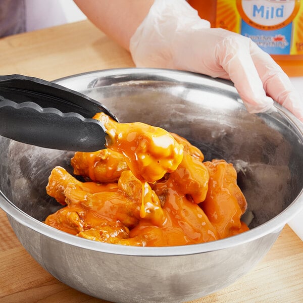 A gloved hand mixes Frank's RedHot Mild Hot Sauce into food in a bowl.