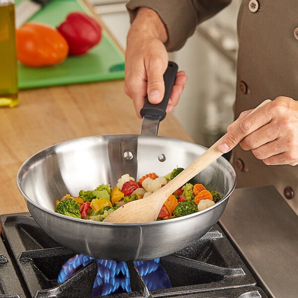 A person cooking vegetables in a Vollrath Tribute stainless steel stir fry pan on a stove.