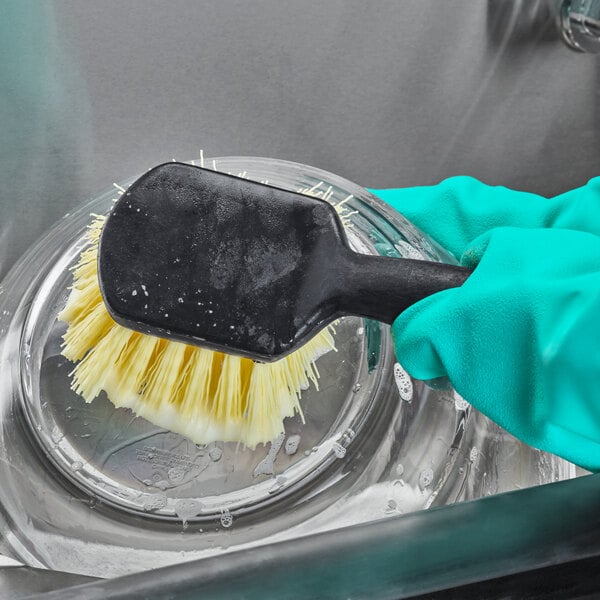 A hand in a glove using a Carlisle Sparta utility brush to clean a dish.