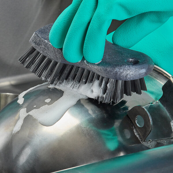 A person wearing gloves cleaning a metal object with a Carlisle Sparta black handheld scrub brush.
