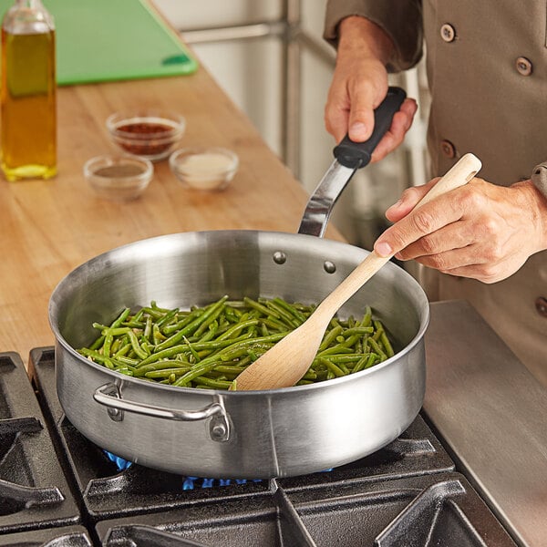 A person sautéing green beans in a Vollrath stainless steel saute pan with a black handle.