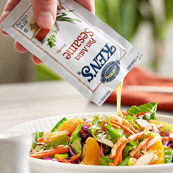 A hand pouring Ken's Foods Pan Asian Sesame Dressing packet over a salad