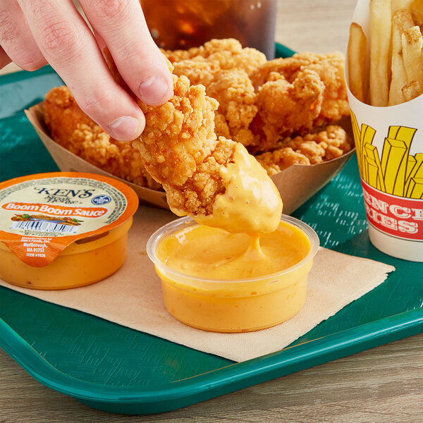 A hand dipping a piece of fried chicken into a container of Ken's Boom Boom Sauce.