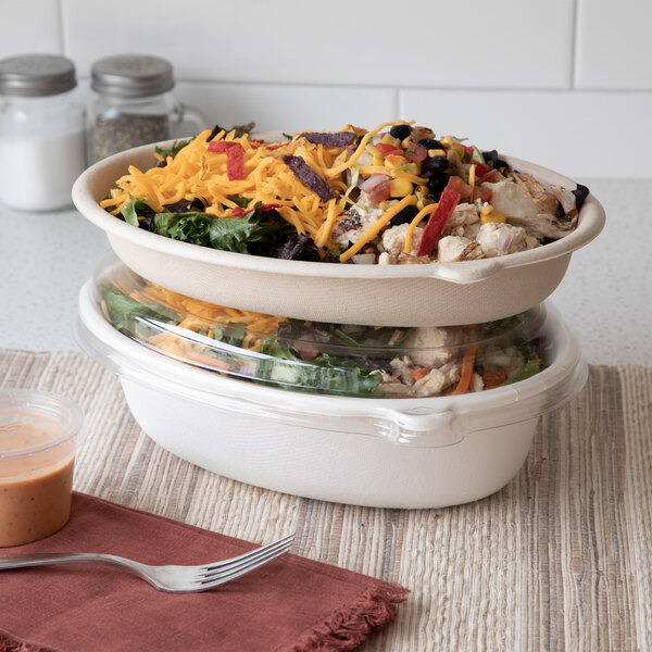 An oval-shaped, compostable sugarcane bowl filled with salad, shown stacked on another bowl with a clear lid.