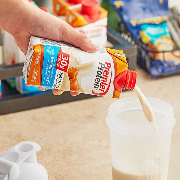 A hand pouring Premier Protein caramel protein shake into a cup.