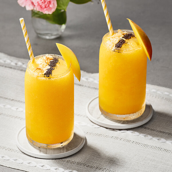 Two glasses of Goya mango smoothie with straws and fruit slices on a table.