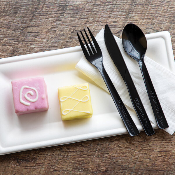 A rectangular white compostable sugarcane plate holding two small decorated cakes, with black plastic cutlery and a napkin beside them.