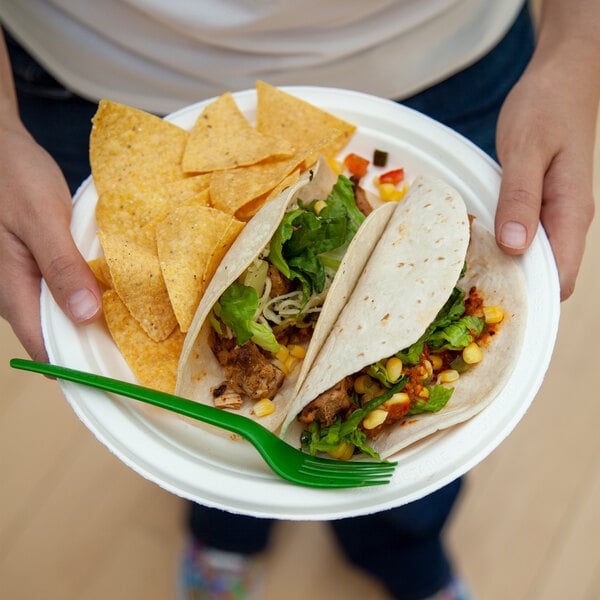 A round compostable sugarcane plate holding two tacos, tortilla chips, and a green plastic fork.