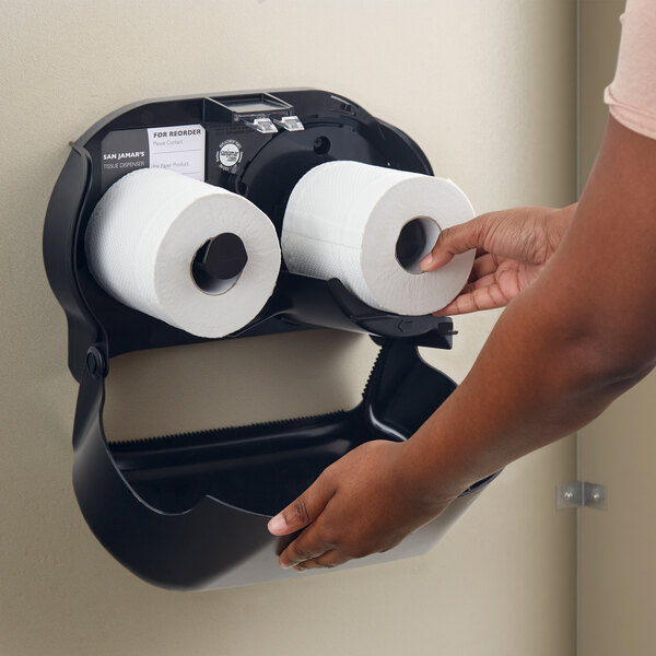 A person putting two rolls of San Jamar toilet paper into a black double roll toilet paper dispenser.