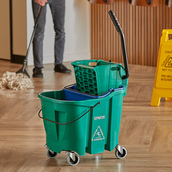 A man in grey pants using a Carlisle OmniFit green mop bucket with wringer on a wood floor.