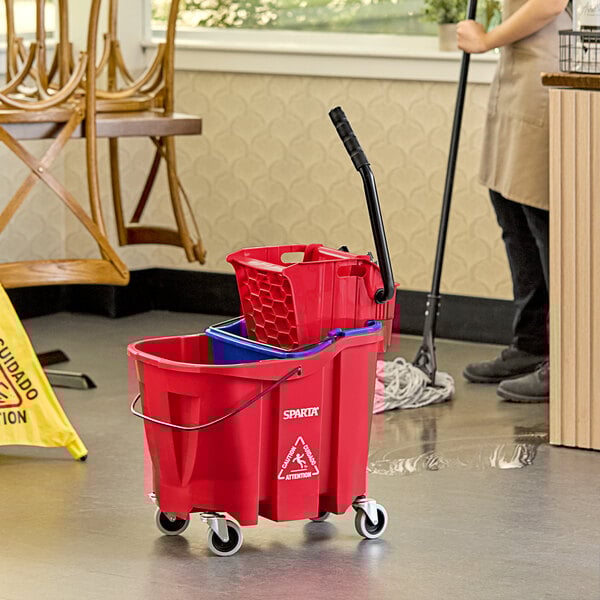 A woman using a Carlisle red mop bucket to clean a hospital cafeteria floor.