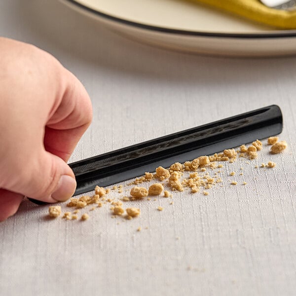 A hand using a Franmara black pocket crumb scraper to clear crumbs from a white tablecloth.
