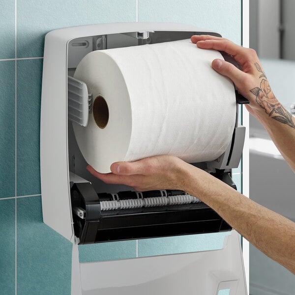 A person loading a large white hardwound paper towel roll into a wall-mounted dispenser.