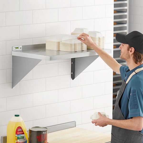 A man in a black cap and apron standing on a Regency stainless steel wall shelf with a stack of plates.