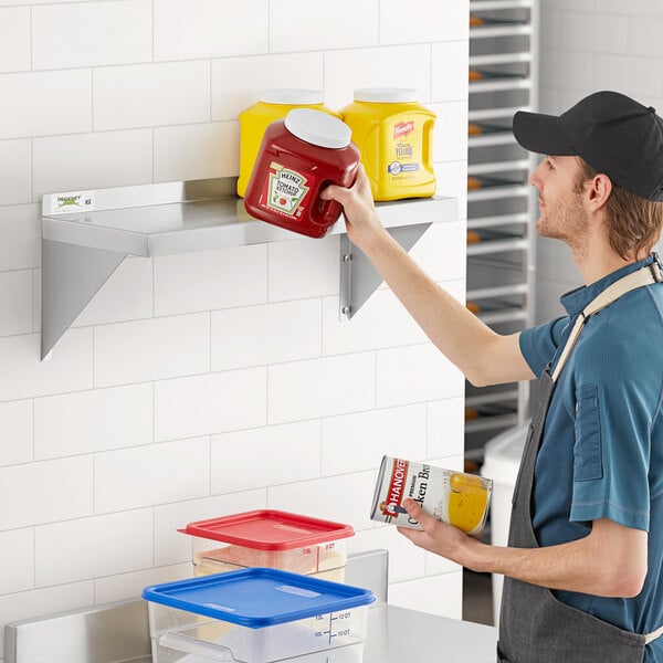 A man standing in front of a Regency stainless steel wall shelf with food including ketchup and chicken broth.