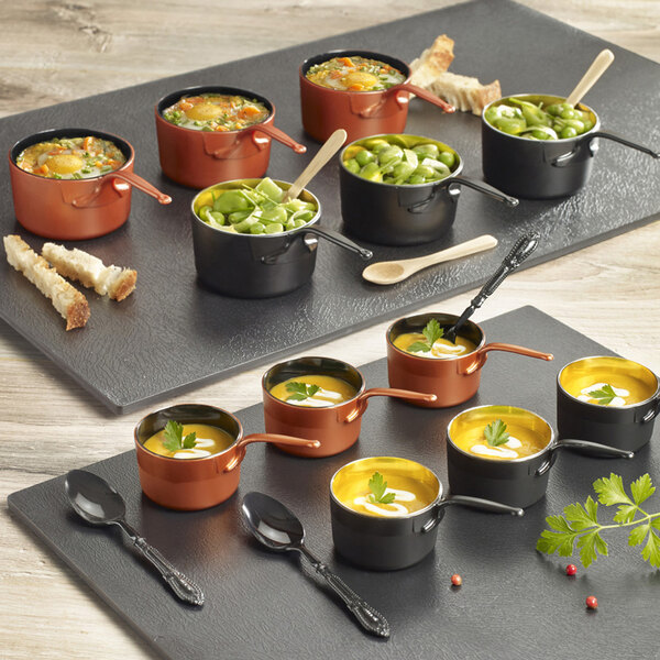 A group of Solia copper and black mini plastic sauce pans filled with soup on a table.