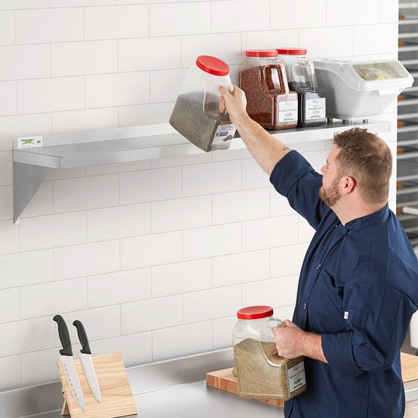 A man standing in front of a Regency stainless steel wall shelf holding a jar of brown sand.