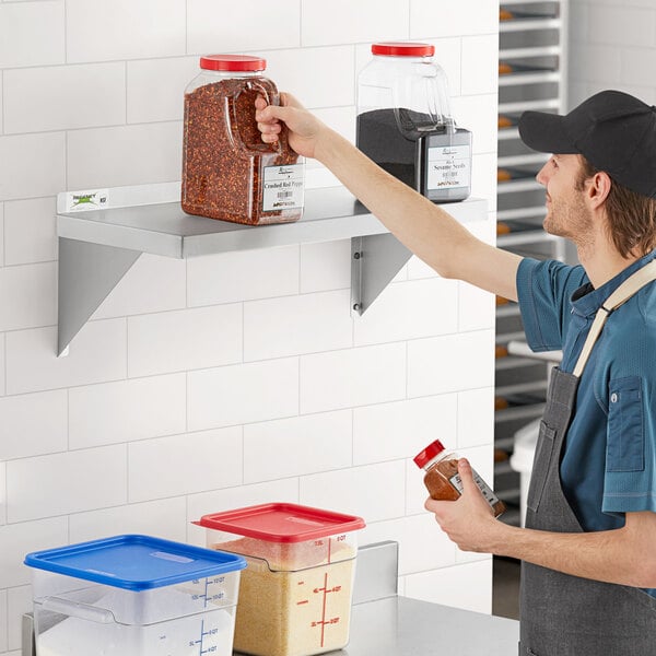 A man in a blue apron places food on a Regency stainless steel wall shelf in a professional kitchen.