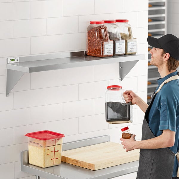 A man in an apron standing in front of a Regency stainless steel wall shelf filled with containers and bottles.