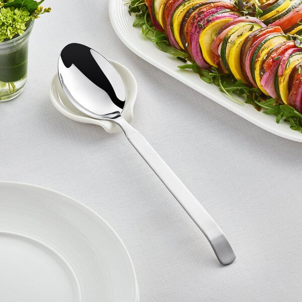 An Acopa stainless steel serving spoon on a white plate next to a plate of vegetables.