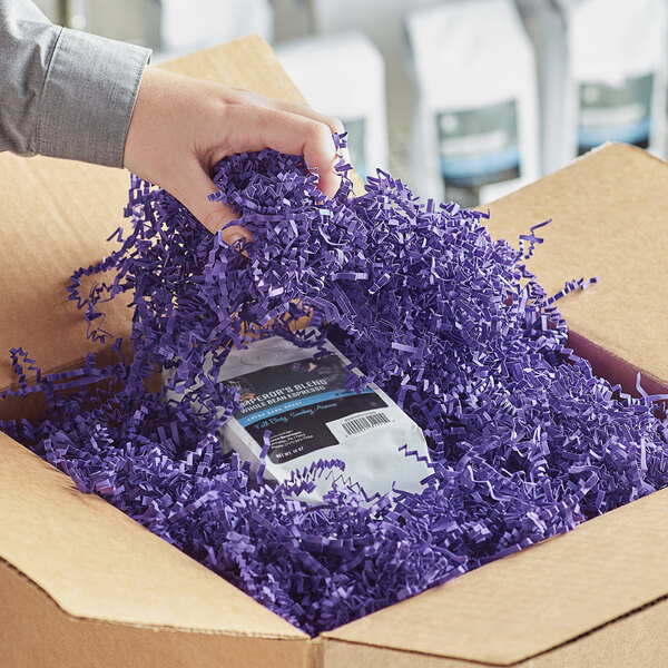 A hand holding a package in a cardboard box filled with Spring-Fill Purple Crinkle Cut paper shred.