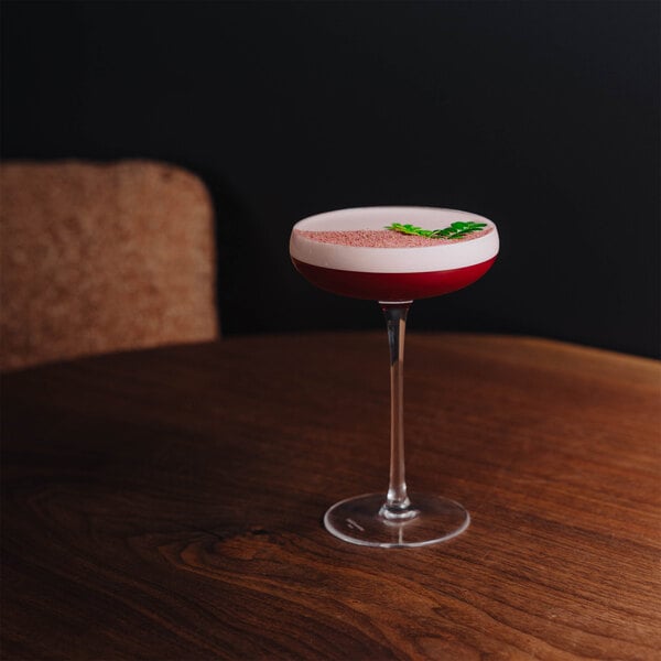 A coupe glass filled with a red cocktail topped with a frothy layer and garnished with a green leaf, placed on a wooden table.