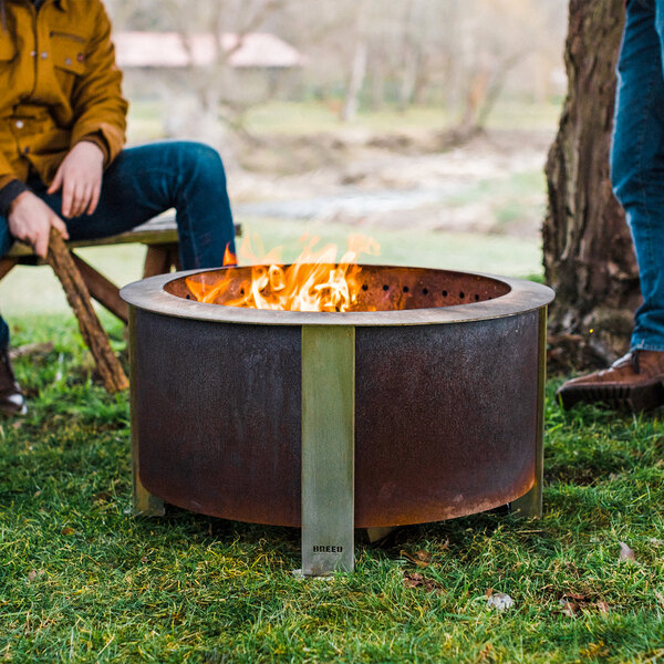 Two men sitting on a bench by a BREEO smokeless fire pit with a fire in it.