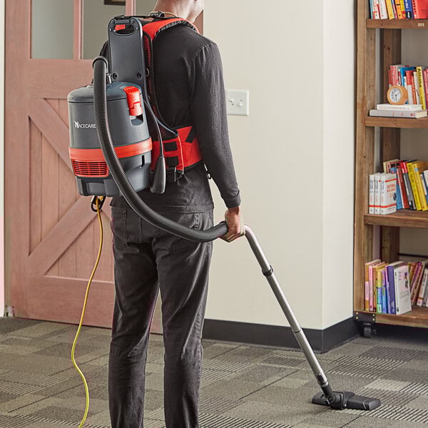 A man wearing a grey and red NaceCare backpack vacuum cleaner vacuuming the floor.