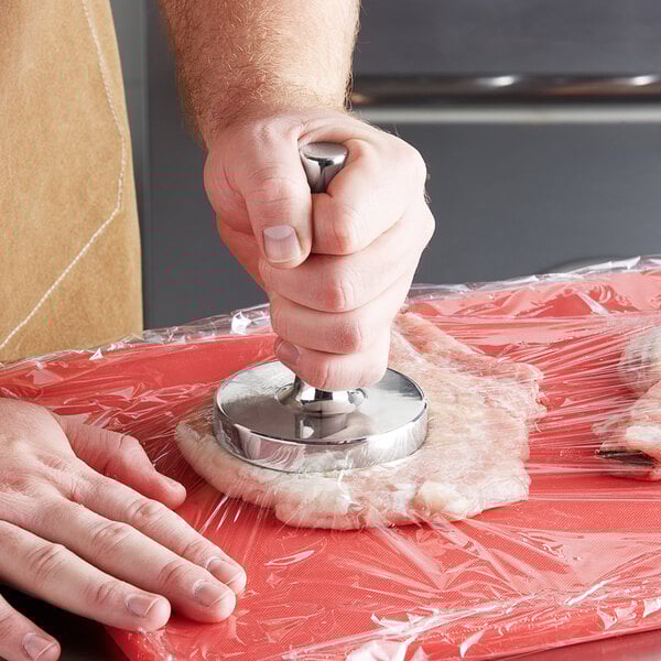 A person using a round metal object to tenderize meat on a counter.