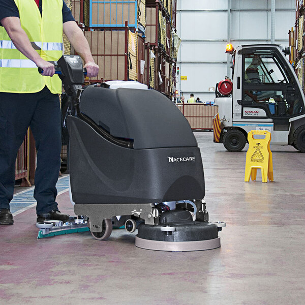 A man using a NaceCare Solutions cordless walk behind floor scrubber with a black cover to clean a warehouse floor.