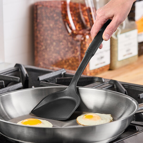 A hand using a Choice black heat-resistant nylon solid turner to cook eggs in a frying pan.