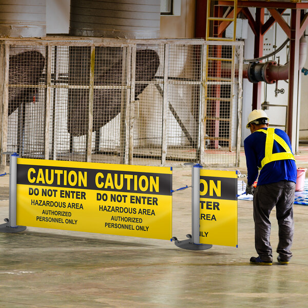 A man in a hard hat and safety vest standing next to a ZonePro single rolling stanchion with a double-sided safety banner with blue accents.