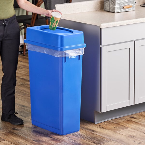 A woman standing next to a Lavex dark blue slim rectangular trash can with a plastic bag inside and throwing a paper cup into it.