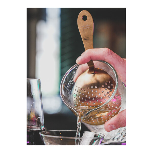 A person using an Arcoroc bronze copper julep strainer to pour liquid into a glass.