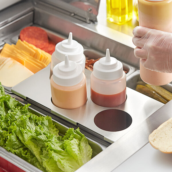 A person using a stainless steel squeeze bottle holder to dispense sauce into a yellow container.