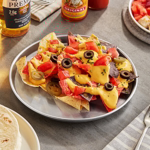 A Choice round aluminum sizzler platter with nachos, cheese, and olives on a table with a bottle of beer.