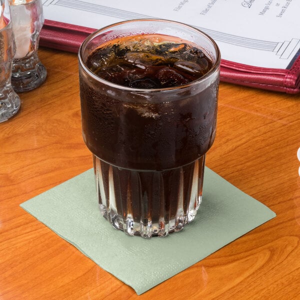 A glass of brown liquid with ice on a soft sage green Hoffmaster cocktail napkin.