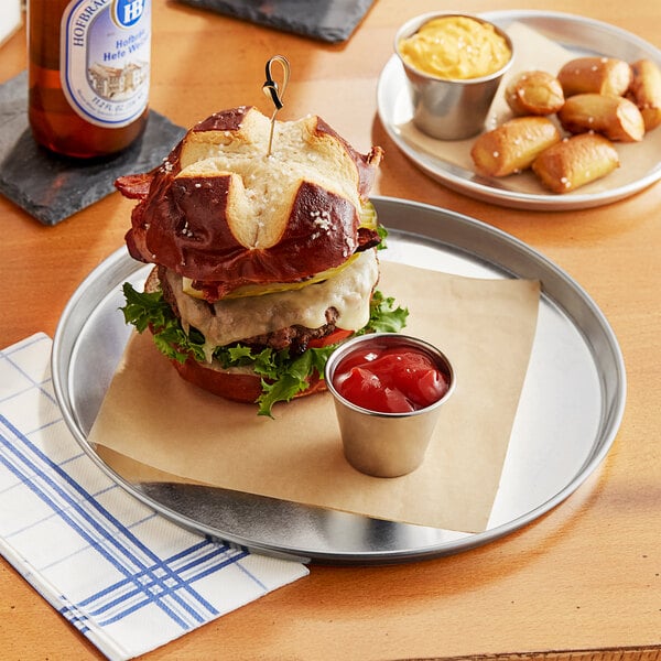 A round aluminum sizzler platter with a large burger and a small cup of red sauce on a table.