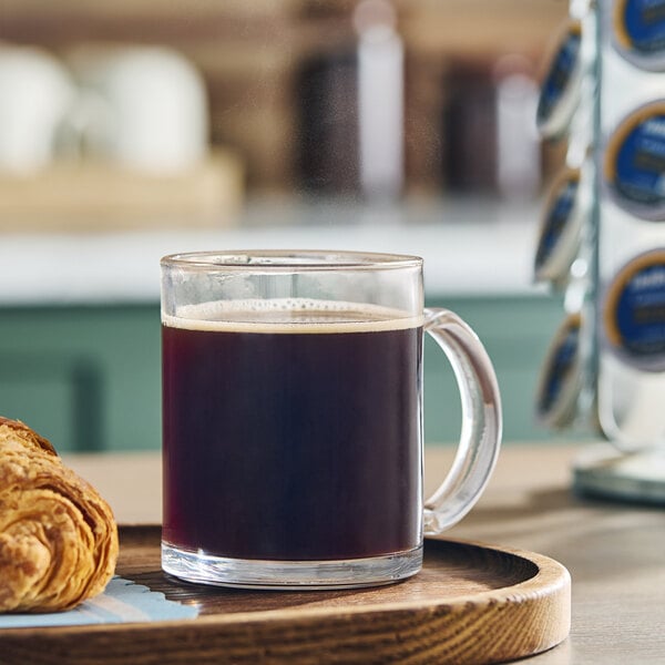 A clear glass mug filled with black coffee sits on a wooden tray next to a croissant, with a rack of coffee pods in the background.