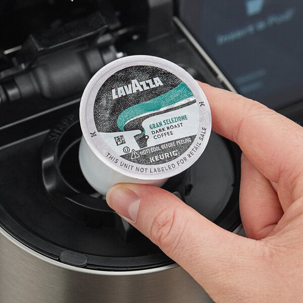 A hand holding a small white and blue round Lavazza K-Cup pod container.