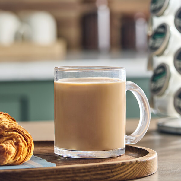 A clear glass mug filled with coffee sits on a wooden tray next to a croissant.
