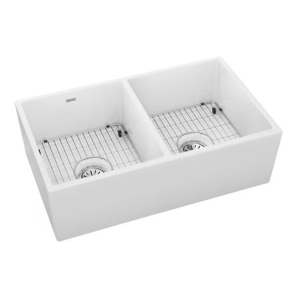 A Zurn Elkay white fireclay double bowl farmhouse sink kit on a kitchen counter with metal racks in each bowl.