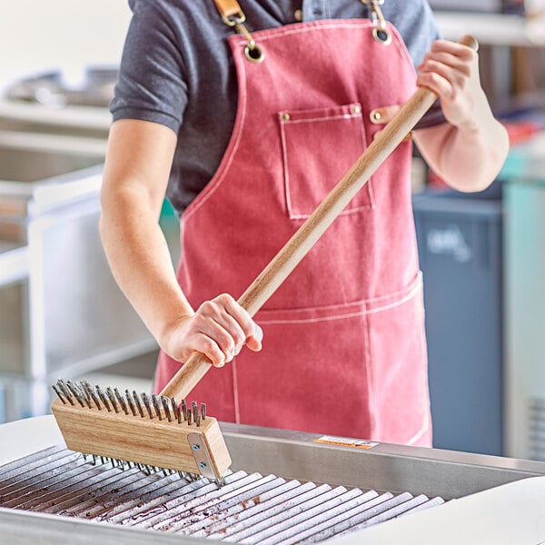 A person in a red apron using a Choice carbon steel bristle grill brush to clean a grill.