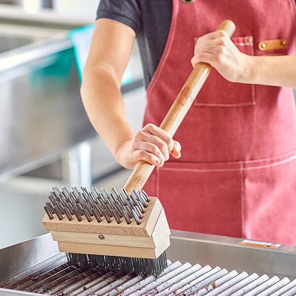 A person in an apron using a Choice steel bristle grill brush to clean a grill.