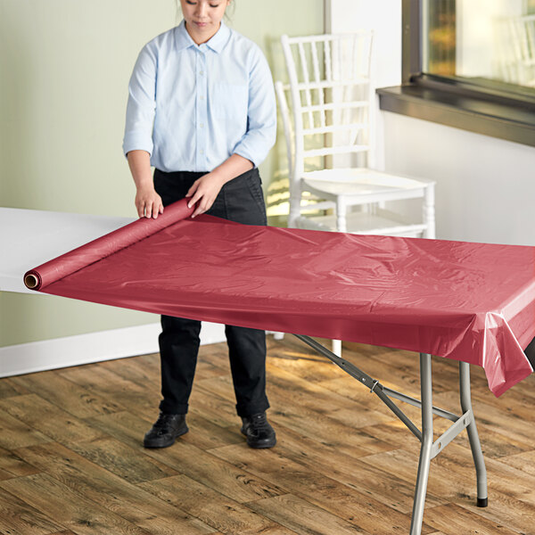 A woman rolling out a burgundy plastic table cover on a table outdoors.