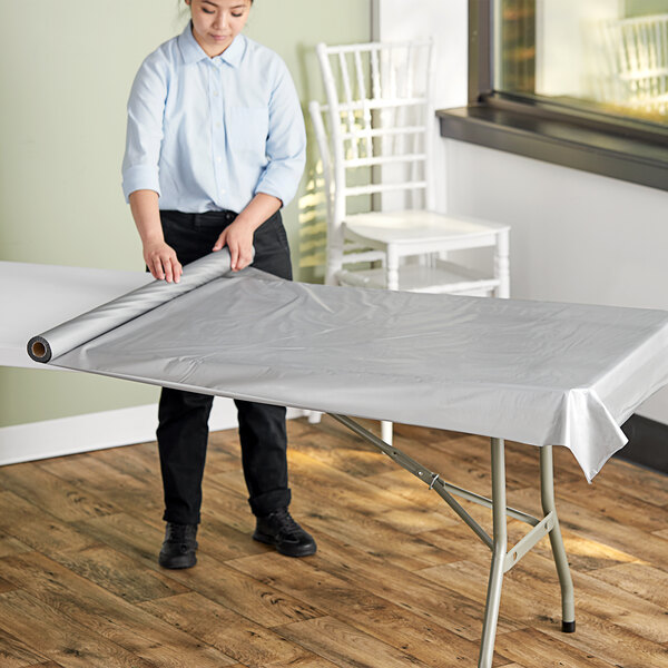 A woman rolling a Table Mate metallic silver plastic table cover onto a table.