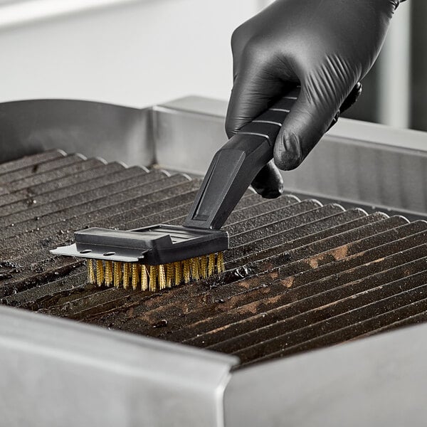 A person wearing black gloves using a Choice brass bristle grill brush to clean a grill.